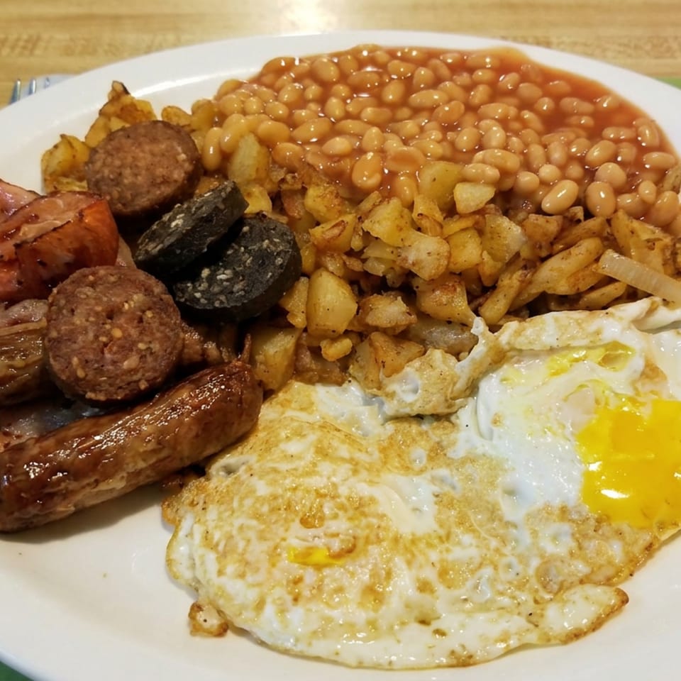 Full English breakfast with fried eggs, sausages, bacon, black pudding, potatoes, and baked beans on a white plate.