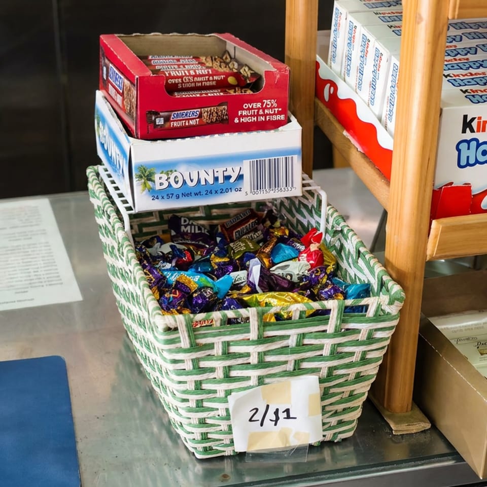 A basket of assorted wrapped candies sits beside boxed chocolates on a wooden display shelf.